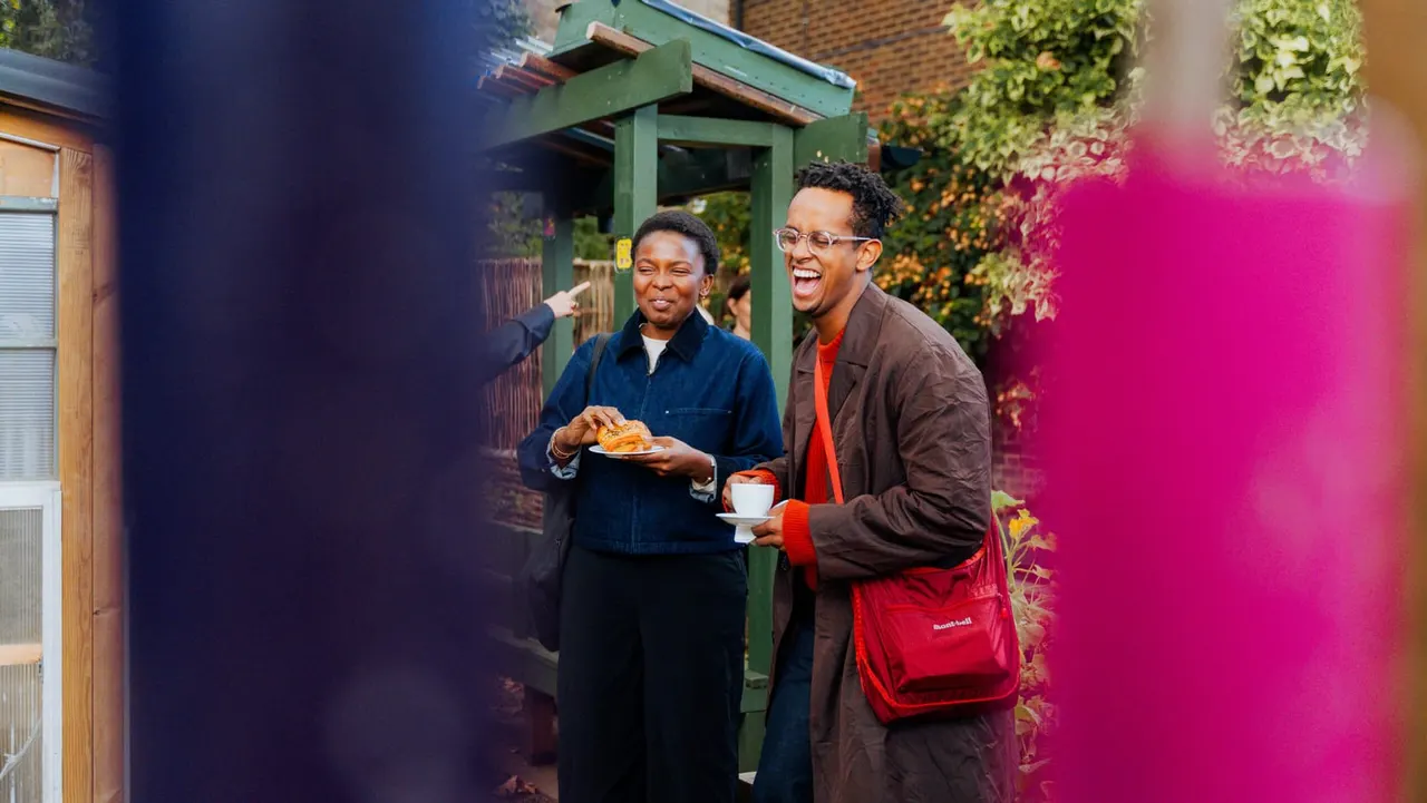 Two people smiling whilst holding food and drinks in a garden courtyard with pink flowering plants and a green pergola.