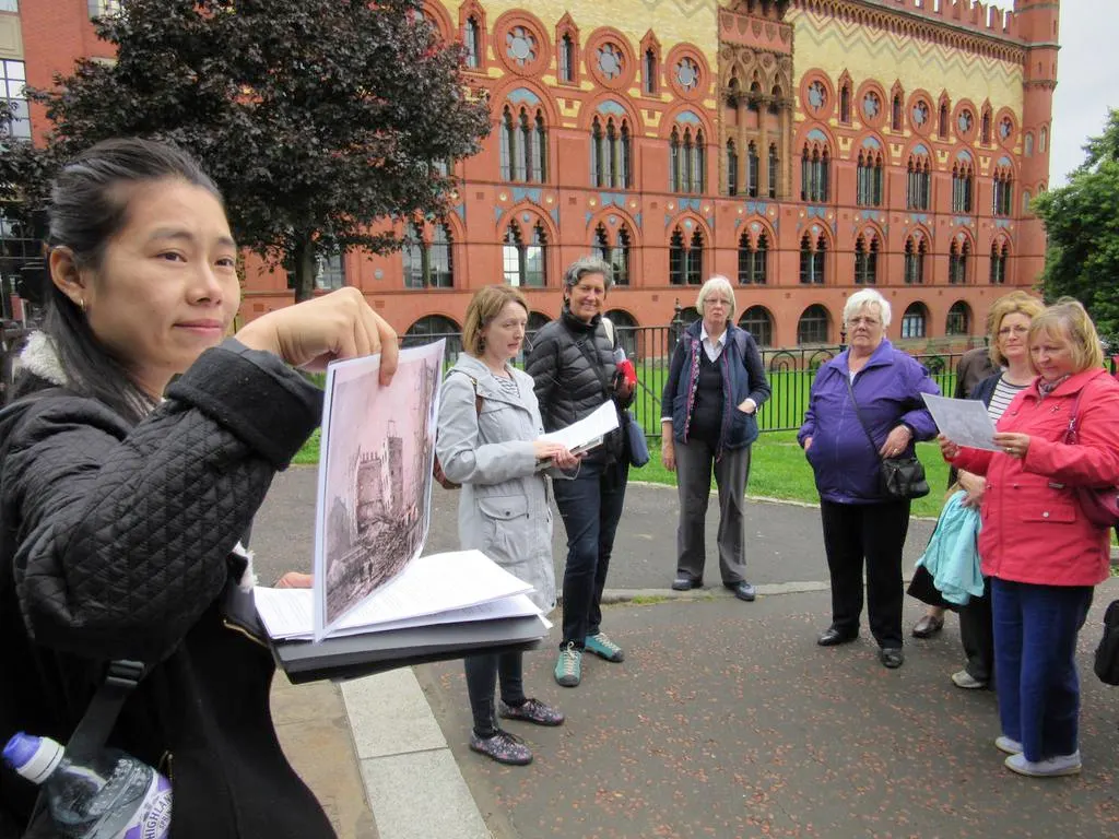 A tour guide holds up a historical image while leading a group of visitors outside a red Victorian building.