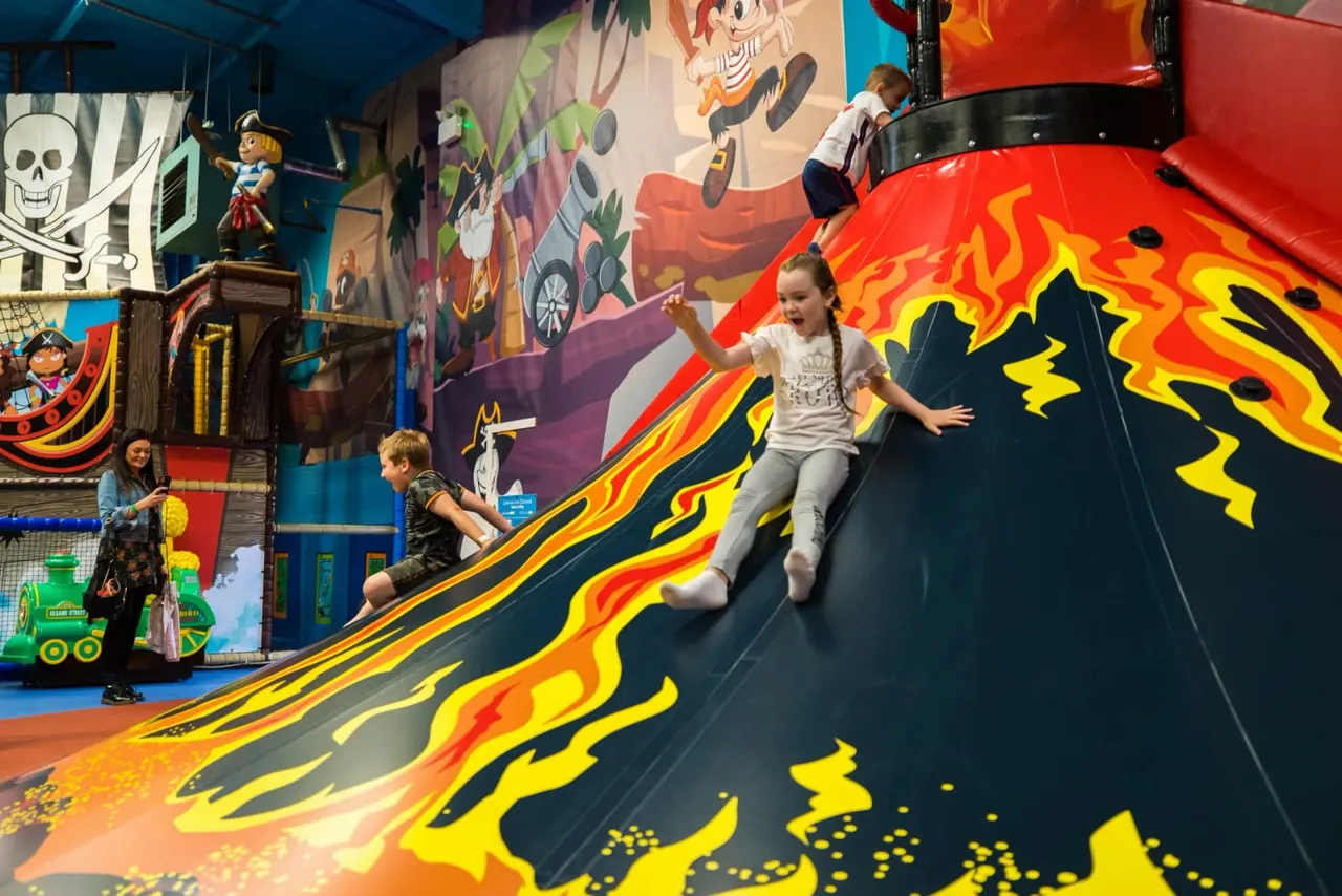 Children sliding down a large indoor slide decorated with flame designs in a soft play adventure area.