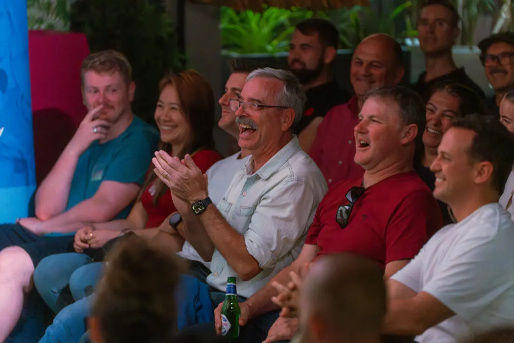 A group of people laughing and cheering together indoors, some holding drinks, in a lively social setting.