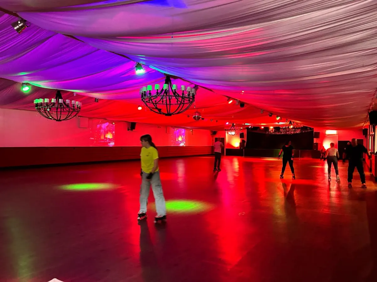 Indoor roller skating rink with neon red and purple lighting, several skaters on the polished floor, and decorative chandeliers overhead.