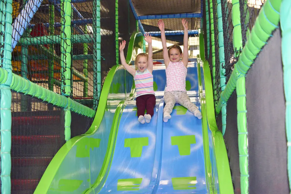 Two young children sliding down a bright green and blue indoor slide with their arms raised.
