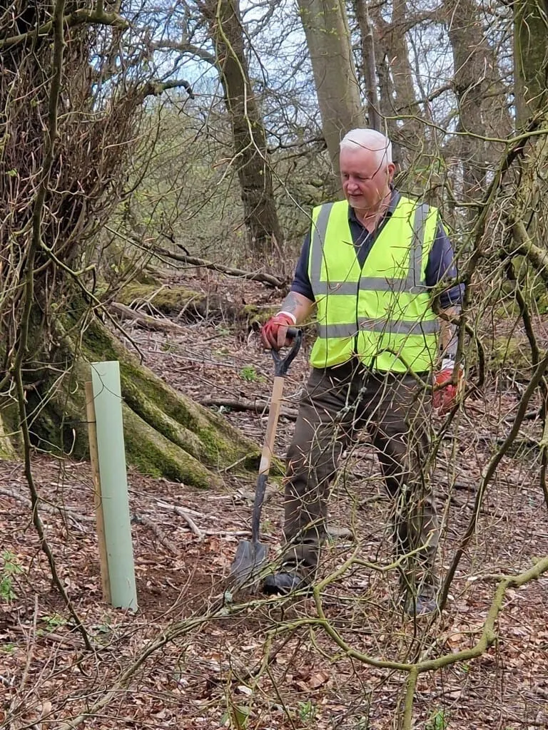 Man in high-visibility vest digging with a spade on a woodland path surrounded by bare branches.