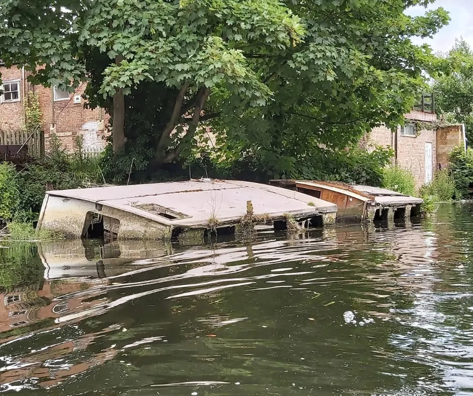 This is a PHOTOGRAPH.

Derelict narrowboat partially submerged in a canal beside brick buildings and overhanging trees.