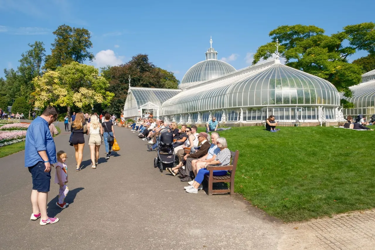 Visitors stroll and sit on benches near a large Victorian glasshouse with distinctive ribbed glass dome in a botanical garden setting.