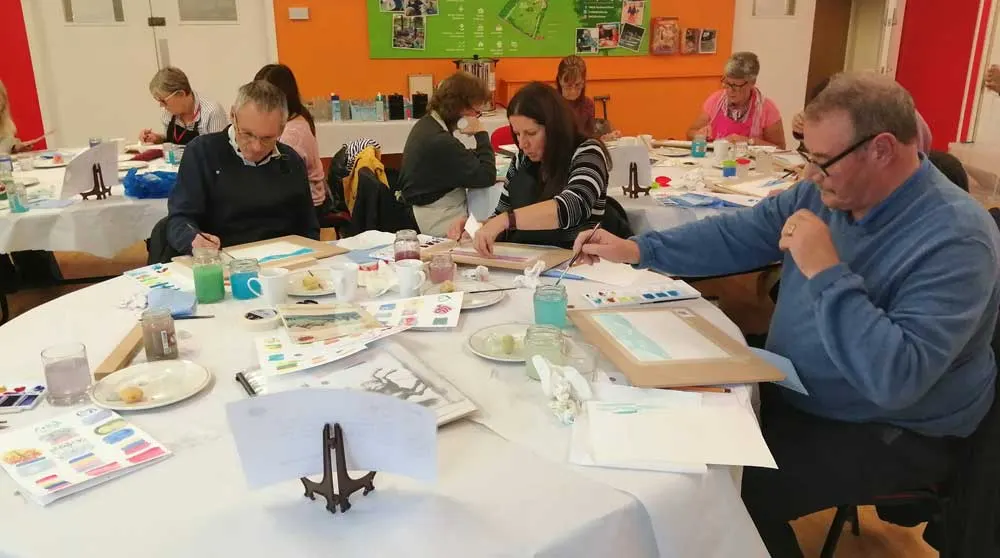 Adults seated at tables engaged in a painting or art workshop activity indoors.