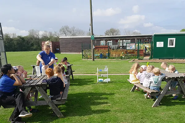 Adults and children seated at picnic tables on a grass field during an outdoor activity session.