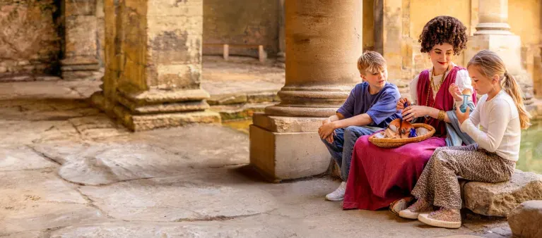 Three children sit together among ancient stone columns at a historic Roman site, sharing food.