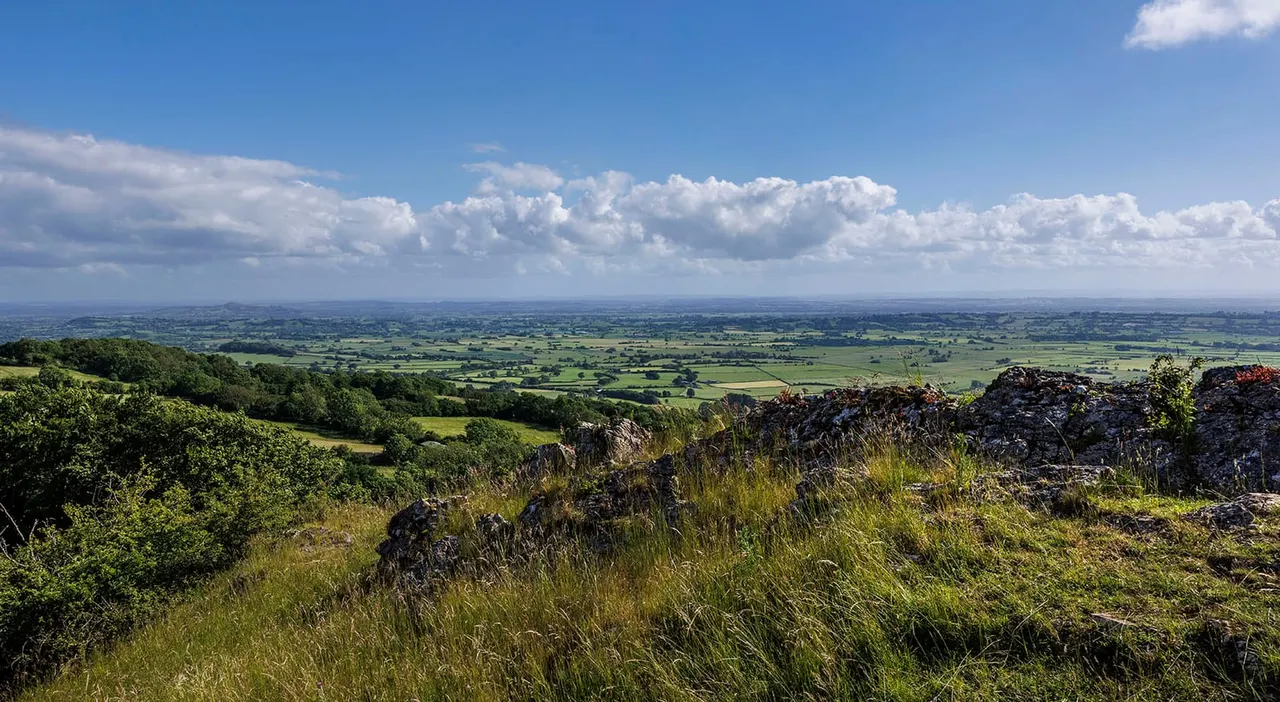 Expansive view across rolling green countryside and patchwork fields from a rocky hilltop under blue sky with white clouds.