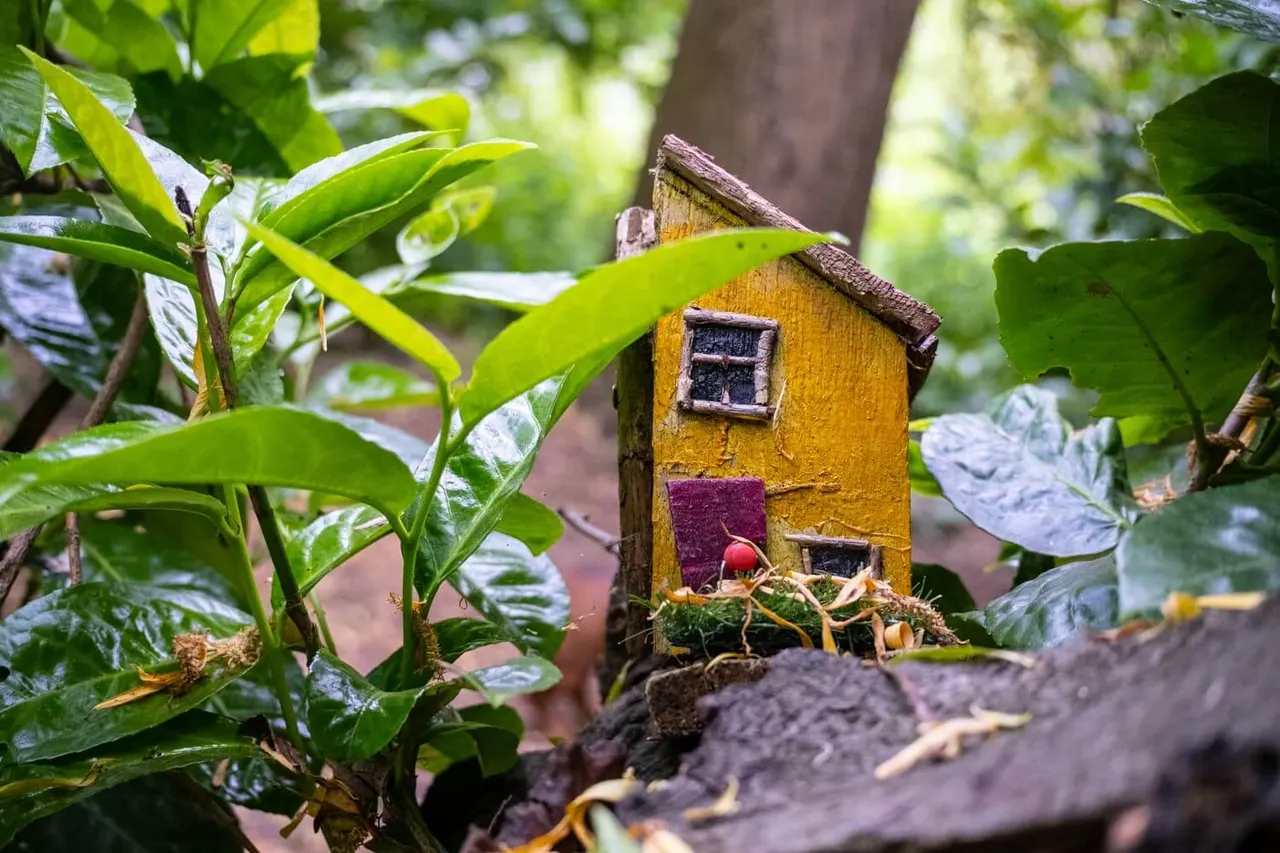 Miniature fairy house with yellow walls nestled among green foliage and tree branches in a woodland garden setting.