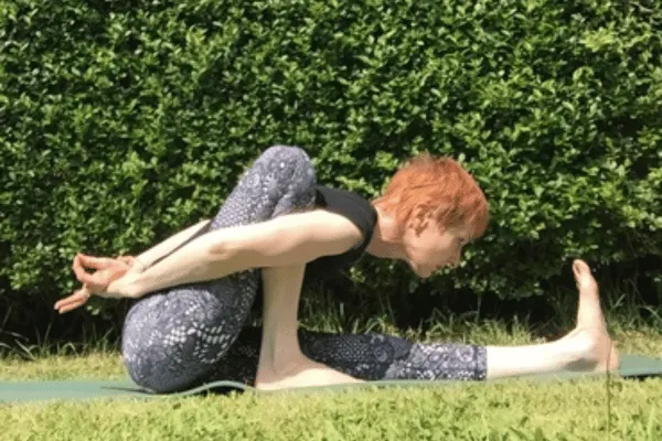 Person performing a seated forward fold stretch on grass against a green hedge backdrop.