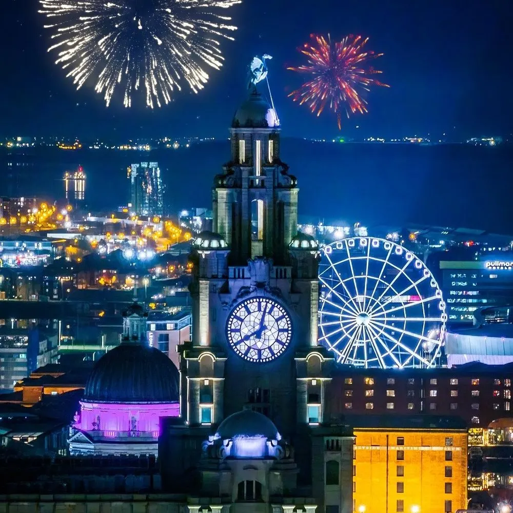 Fireworks explode above the illuminated Liver Building and Wheel of Liverpool at night, with the city lit up below.