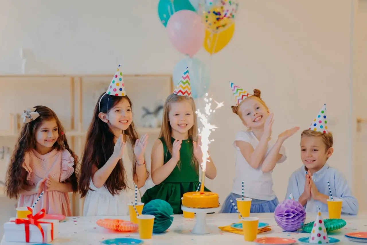 Five children wearing party hats celebrate around a birthday cake with sparklers and balloons at a party table.