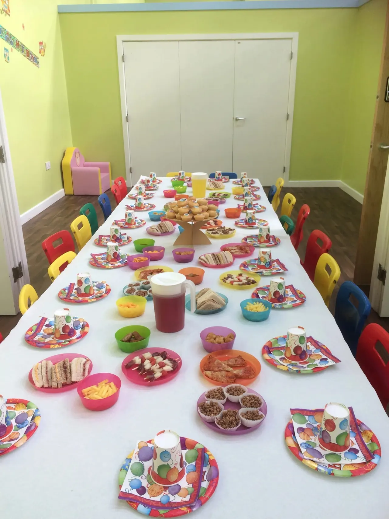 A bright children's party room with a long table set with colorful plates, food, and drinks, surrounded by small coloured chairs.