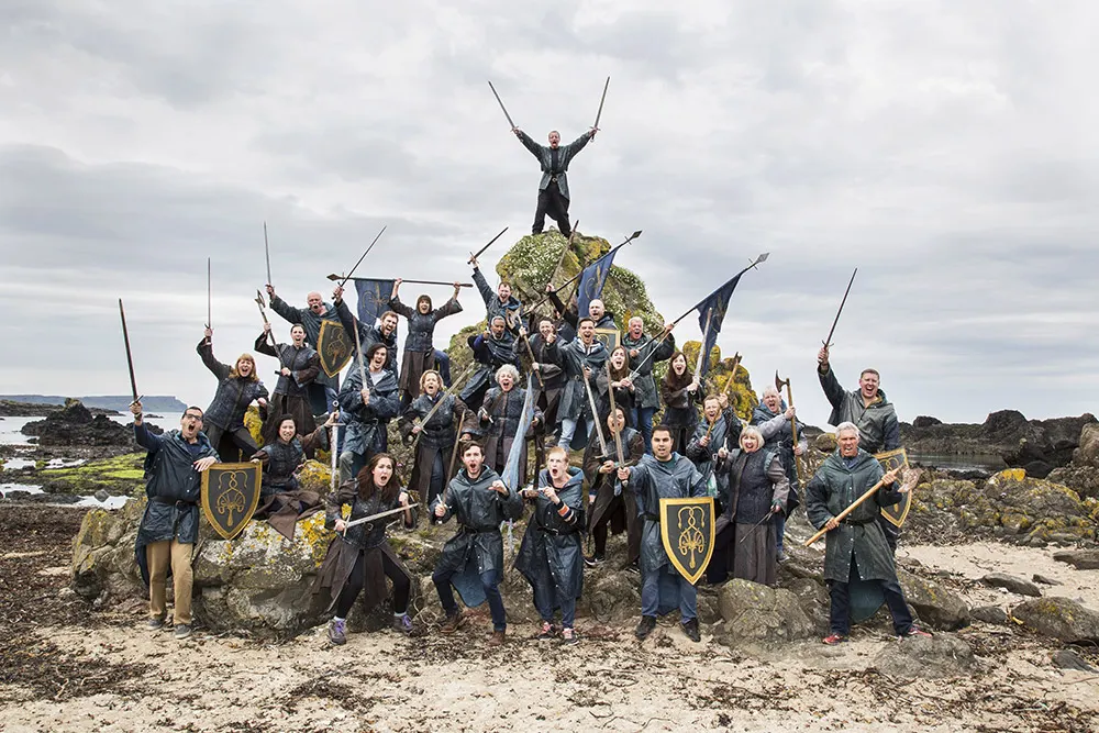 Large group in medieval costume and armour posing with weapons and flags on a rocky coastal beach.