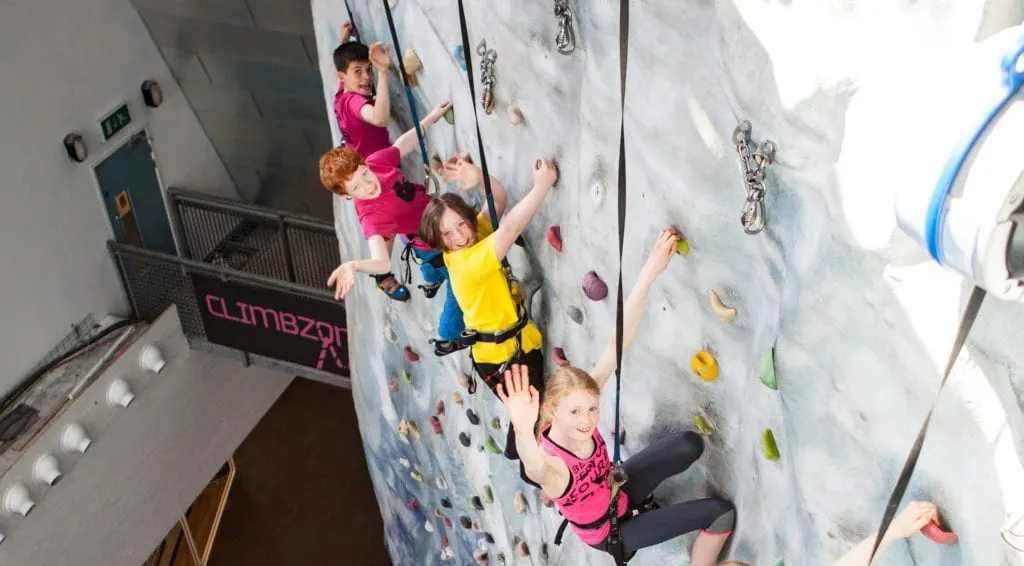Three children climbing an indoor rock wall with safety harnesses at Climbzone.