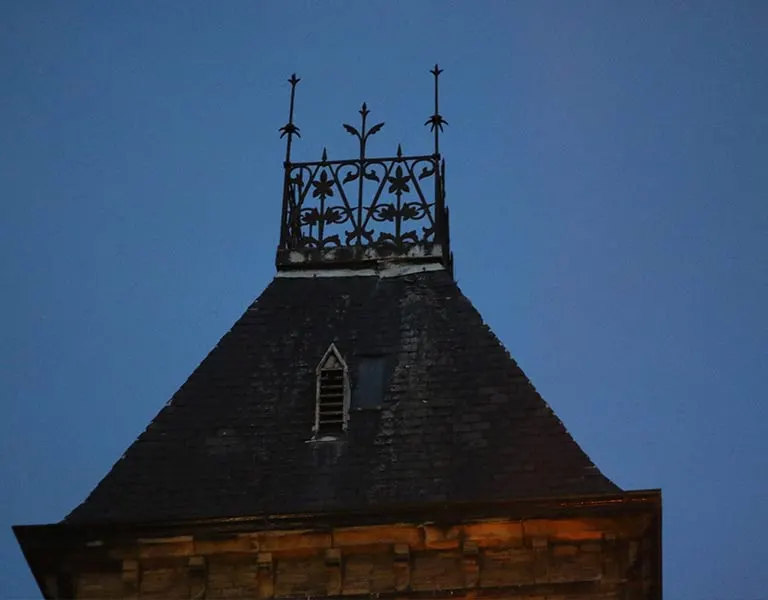 Ornate wrought iron weathervane and chimney silhouetted against a clear blue sky at dusk.