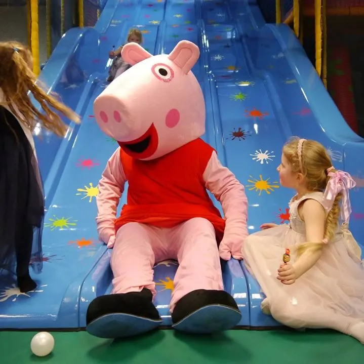 A costumed pink character in a red shirt sits at the bottom of a blue inflatable slide whilst a young girl in a dress watches nearby.