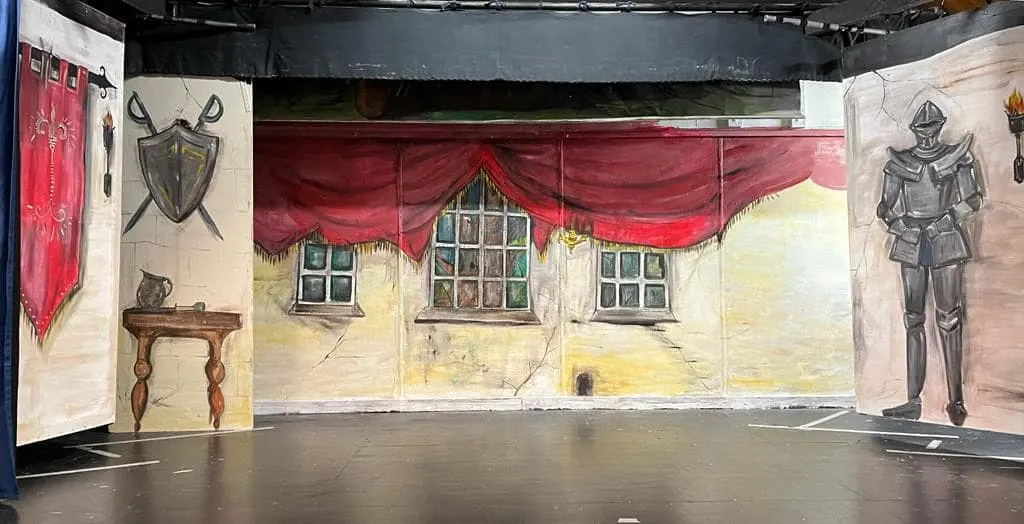 Theatre stage set with medieval-themed painted backdrops featuring shields, armour, red drapes and castle windows.