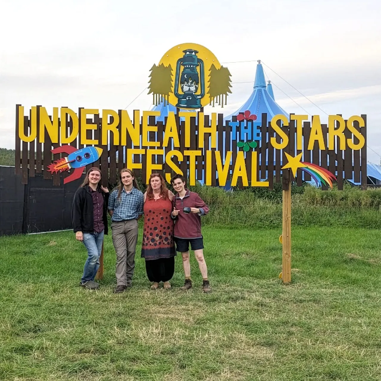 Four people standing on grass in front of a large colourful festival sign with a rocket and stars design.