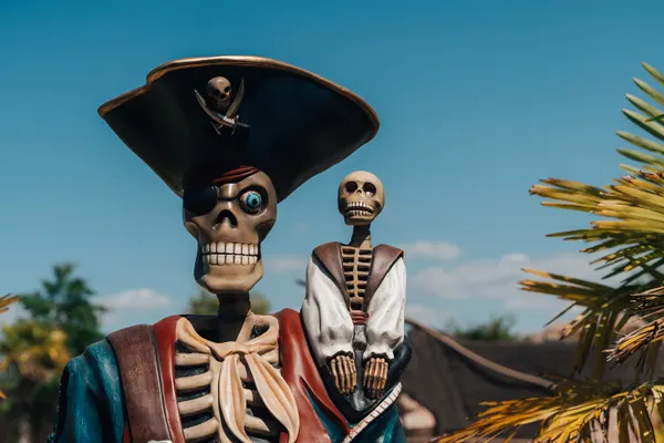 Two skeleton figures dressed as pirates with a tricorn hat and period costume against a blue sky and tropical foliage.