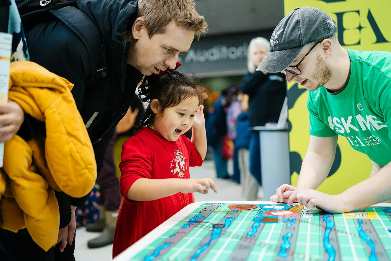 Young child in red shirt engaging with a staff member at an interactive activity table whilst adults supervise nearby.
