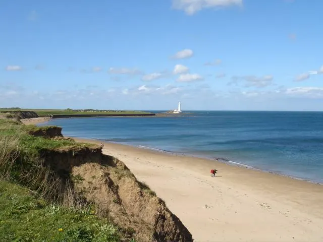 Coastal view of a sandy beach with grassy cliffs, a lone person in the distance, and a lighthouse on the headland.
