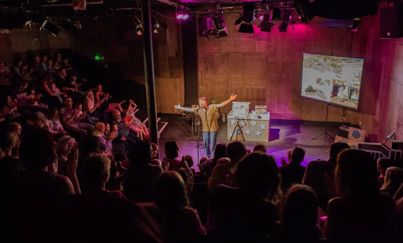 A performer in a yellow striped shirt addresses an engaged audience in a dimly lit theatre with pink and purple stage lighting.