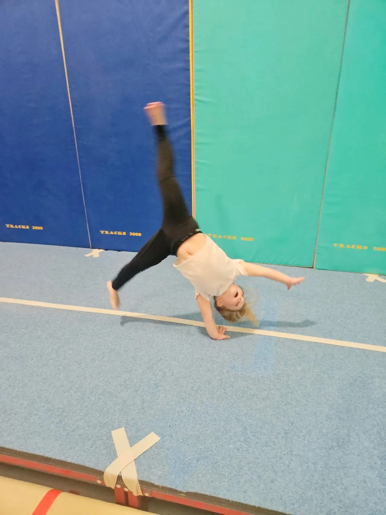 Young person performing a handstand on a blue gymnasium mat against coloured wall panels.