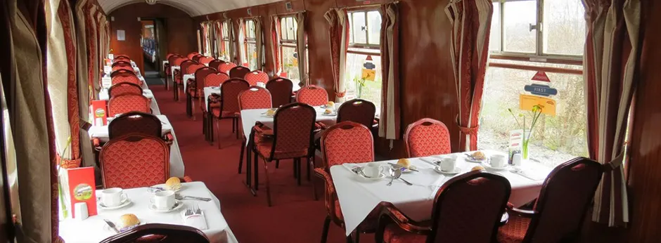Interior of a heritage railway dining car with red upholstered chairs, white-clothed tables, and large windows overlooking countryside.