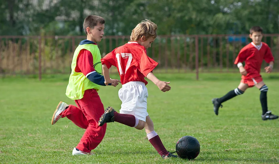 Young children playing football match on grass pitch, one in red kit with ball, one in high-visibility bib defending.