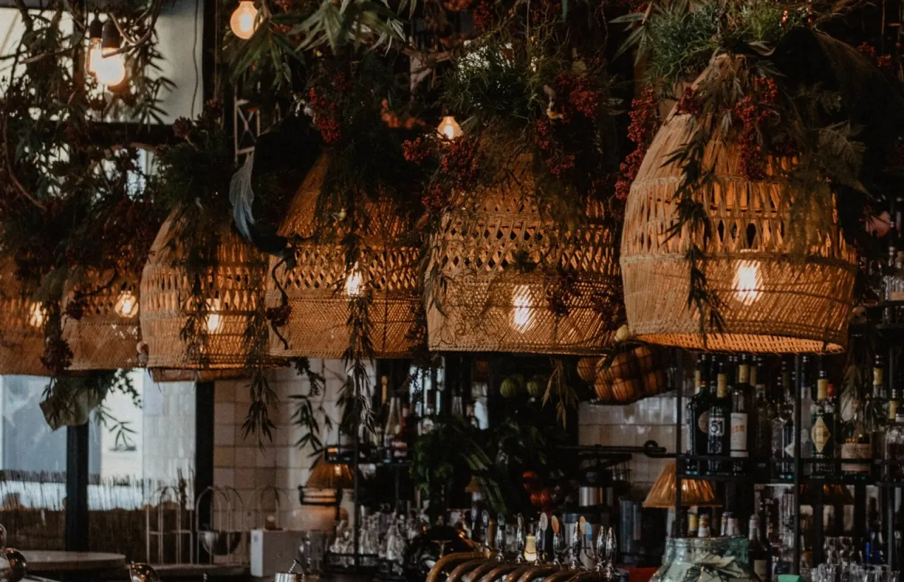 Woven pendant lights and cascading foliage with red berries hang above a bar interior with bottles and glassware.