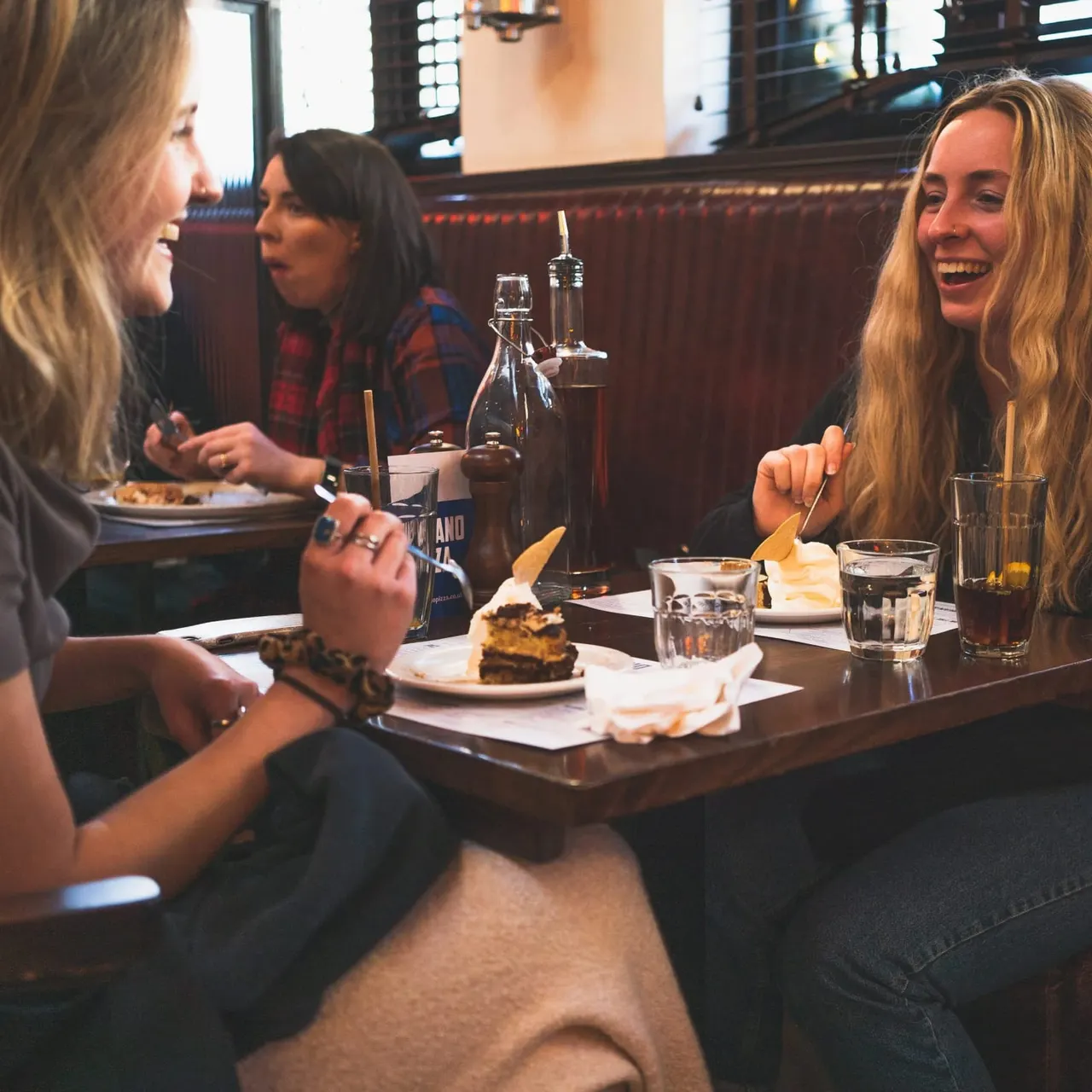 Three women dining and laughing together in a booth at a restaurant with food and drinks on the table.