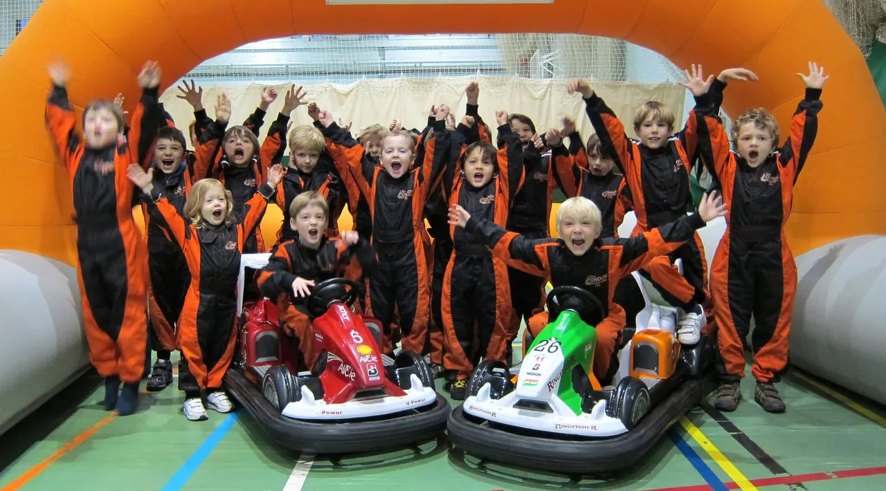 Group of children in orange and black racing suits celebrating with bumper cars in an indoor karting arena.