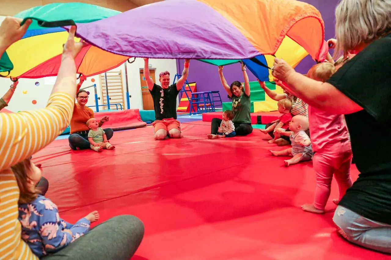Adults and toddlers playing together under a colourful parachute on a red padded floor in an indoor play area.