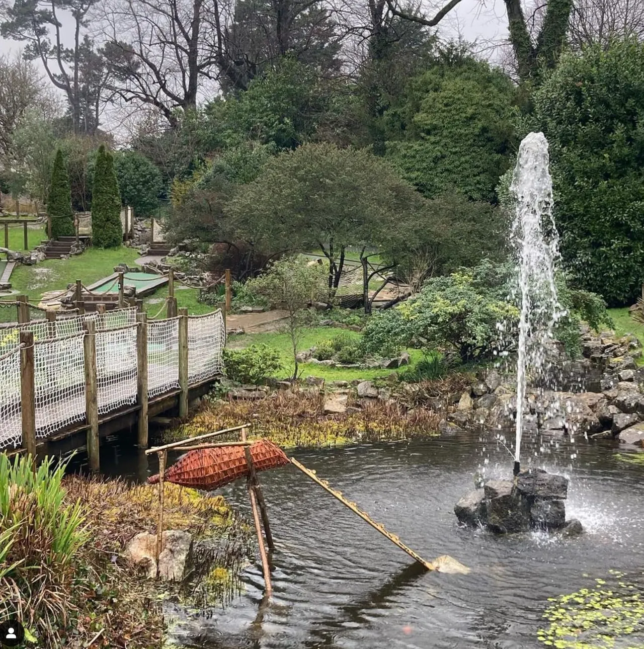 A water fountain sprays beside a pond with a red boat and wooden bridge in a landscaped garden with trees.