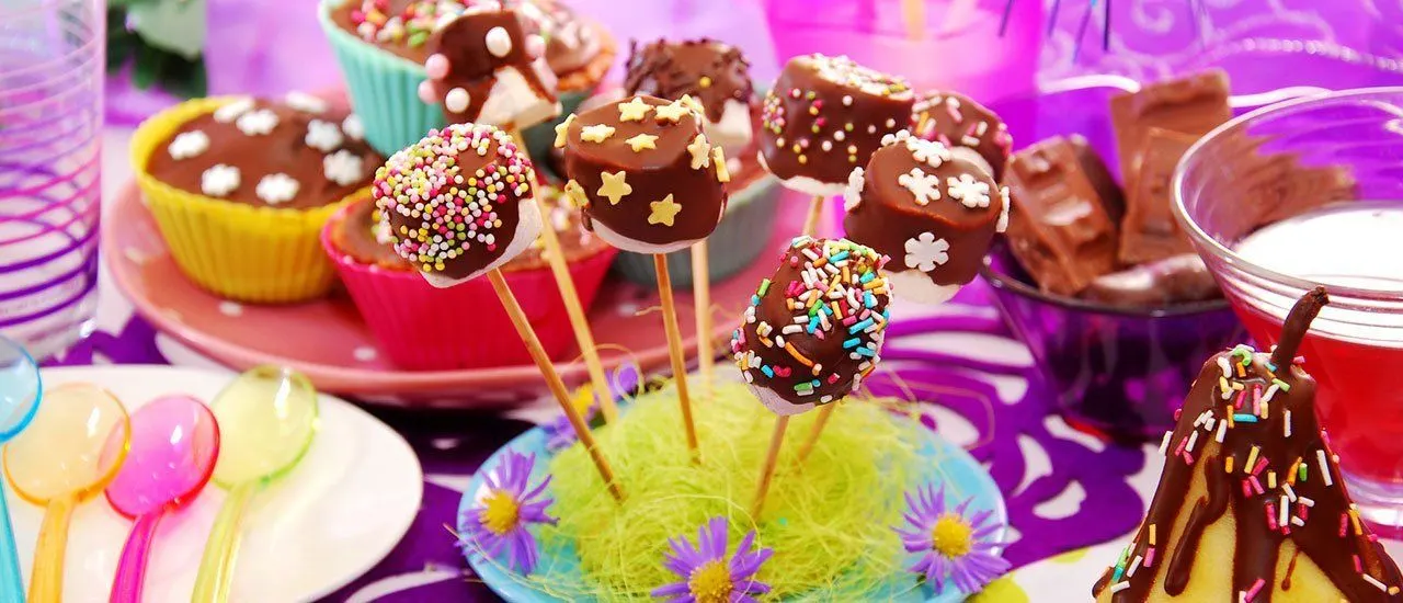 Colourful children's party table with chocolate cake pops decorated with sprinkles and sweets, cupcakes, and colourful tableware against a bright purple background.