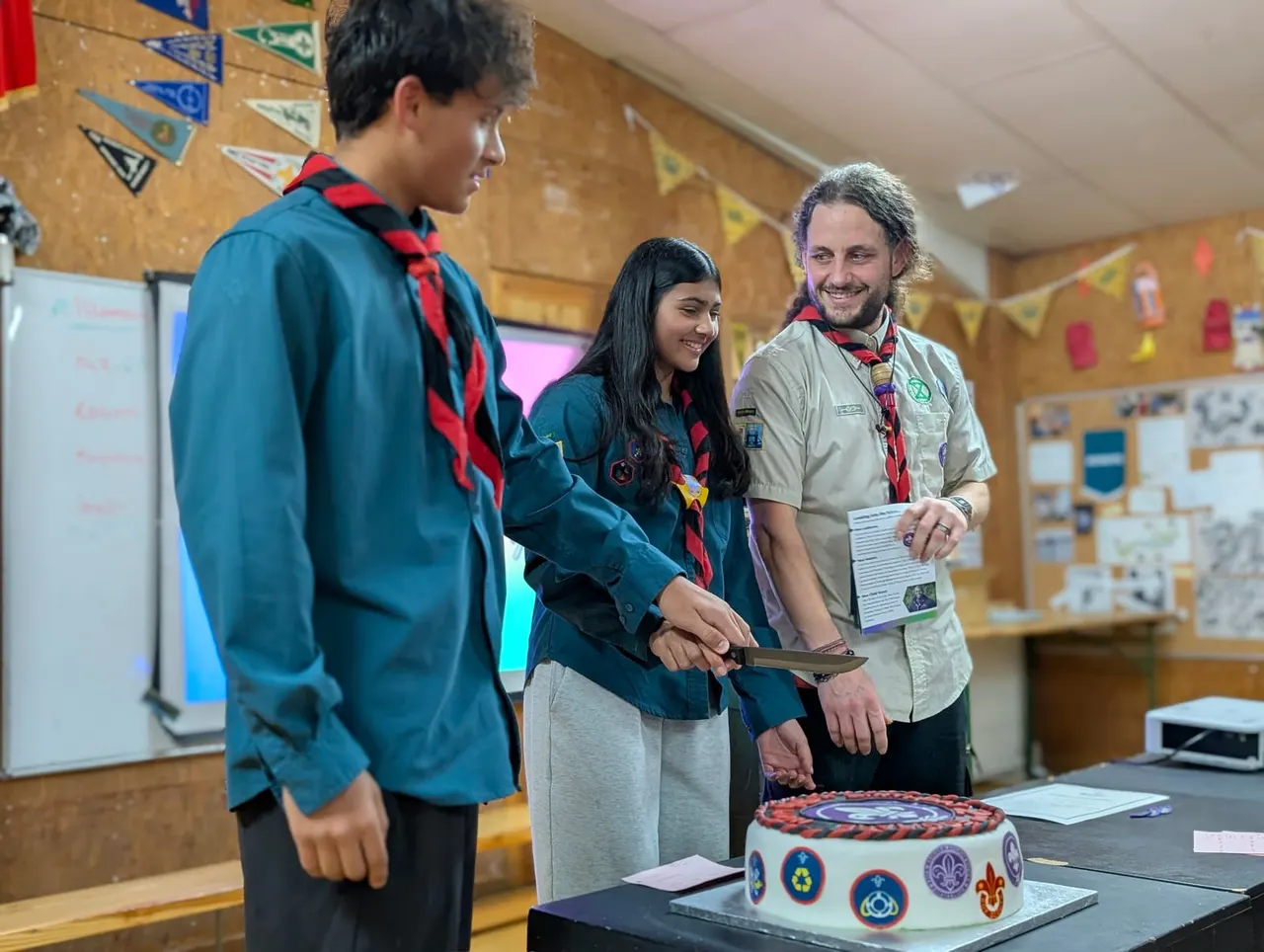 An adult scout leader smiles whilst presenting a decorated cake to two young scouts in uniform inside a scout hall decorated with bunting.