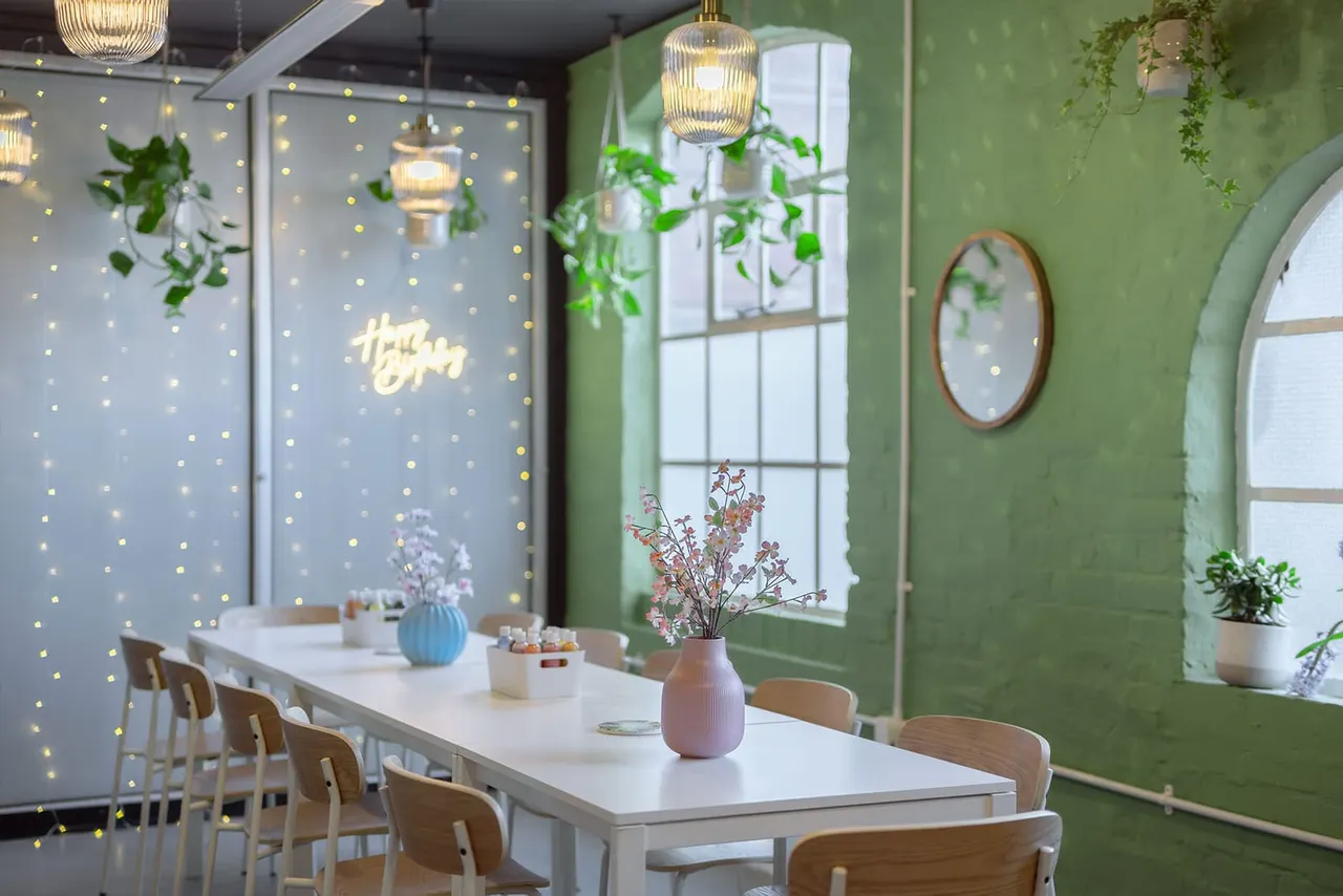 Modern bright café interior with long white table, wooden chairs, hanging plants, pendant lights and green-painted walls.