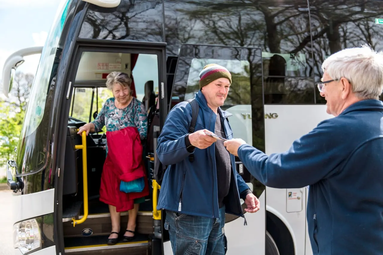Tour guide handing tickets to a passenger boarding a white coach bus, with another passenger visible in the doorway.