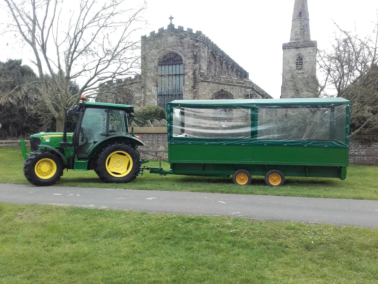 Green and yellow tractor with enclosed green trailer parked on grass, historic stone church buildings in background.