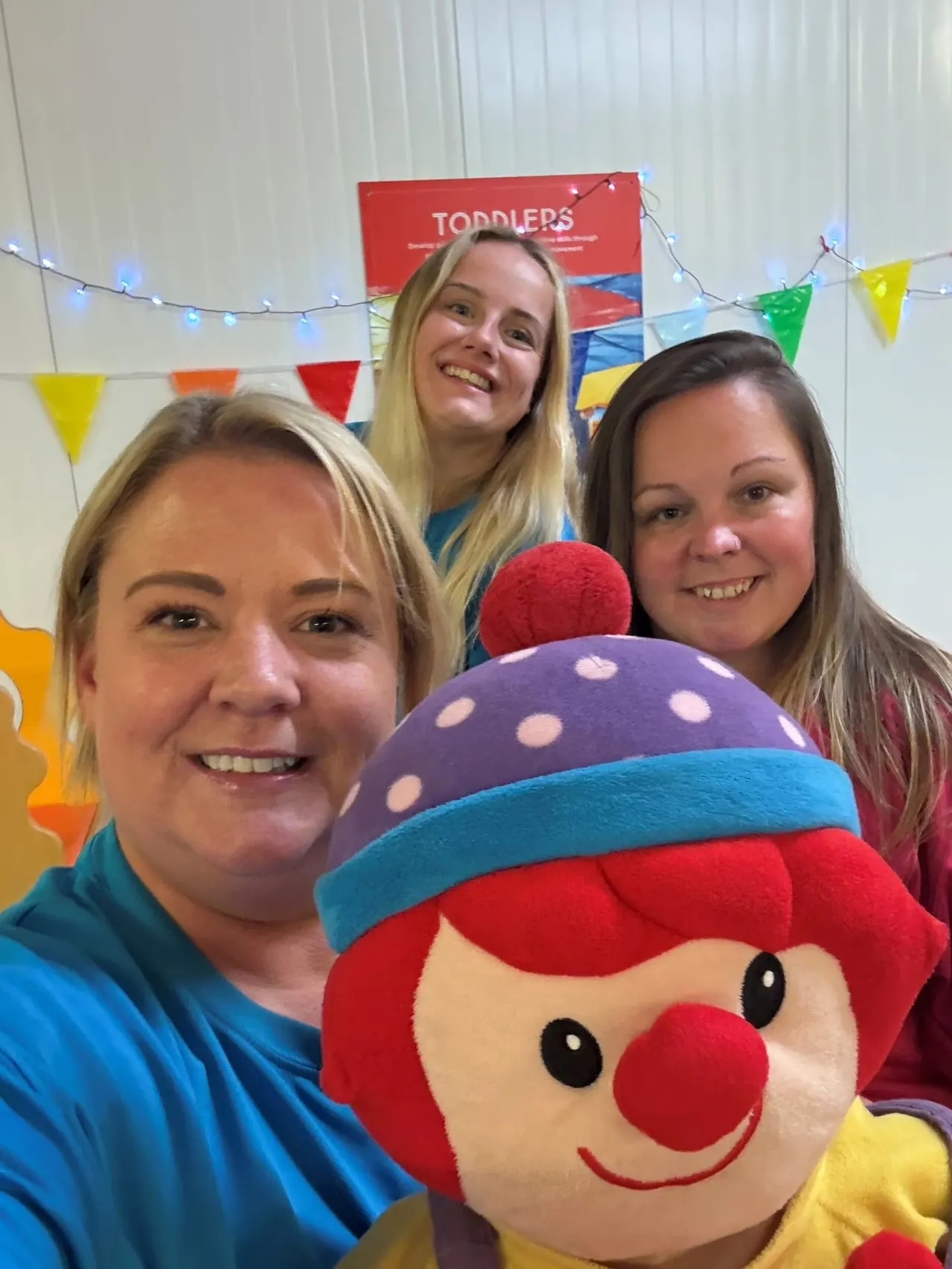 Three smiling adults posing with a large colourful clown character at a decorated indoor play venue.