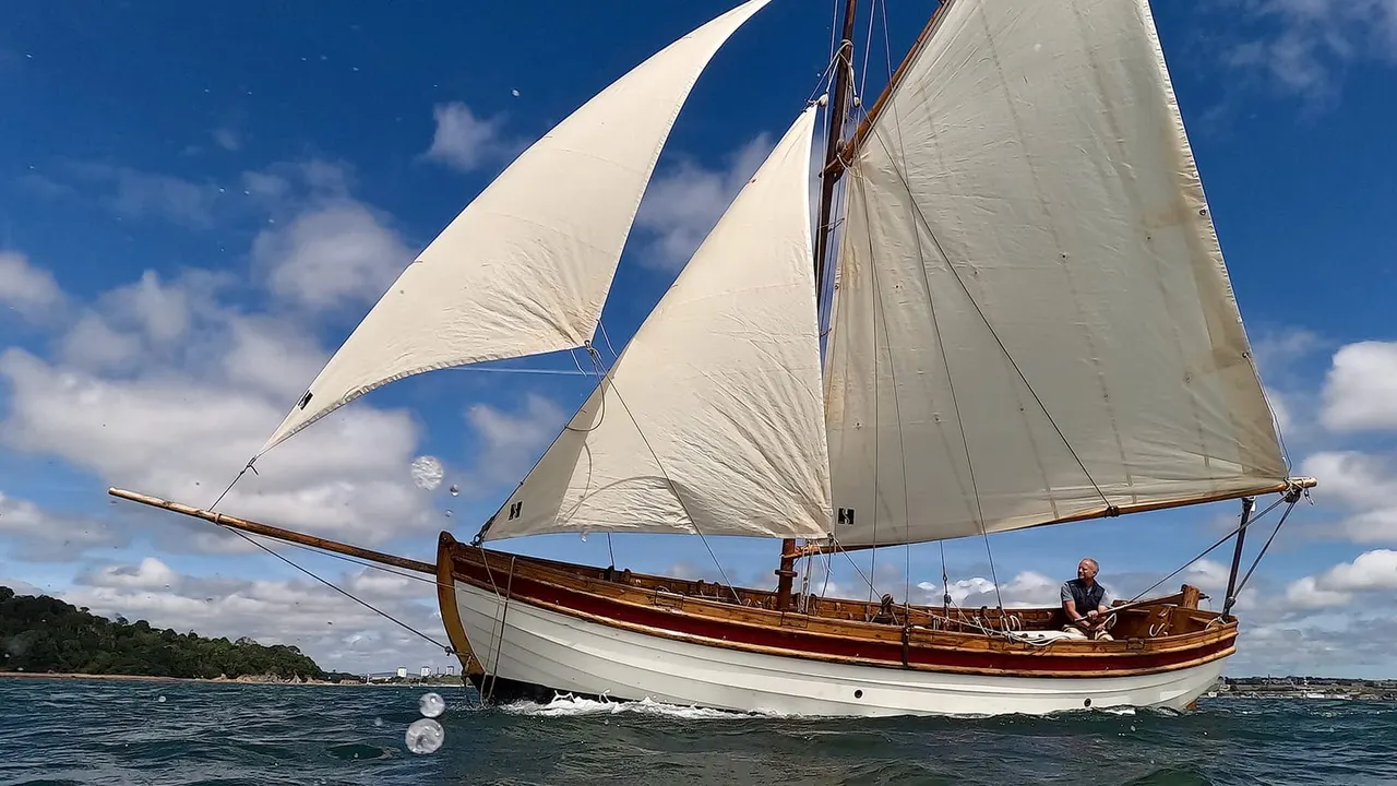 Traditional sailing vessel with cream sails and wooden hull under full sail on blue water with one person visible on deck.