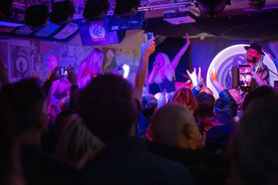 Crowded nightclub dance floor bathed in purple and blue lighting, with people dancing and holding phones aloft.
