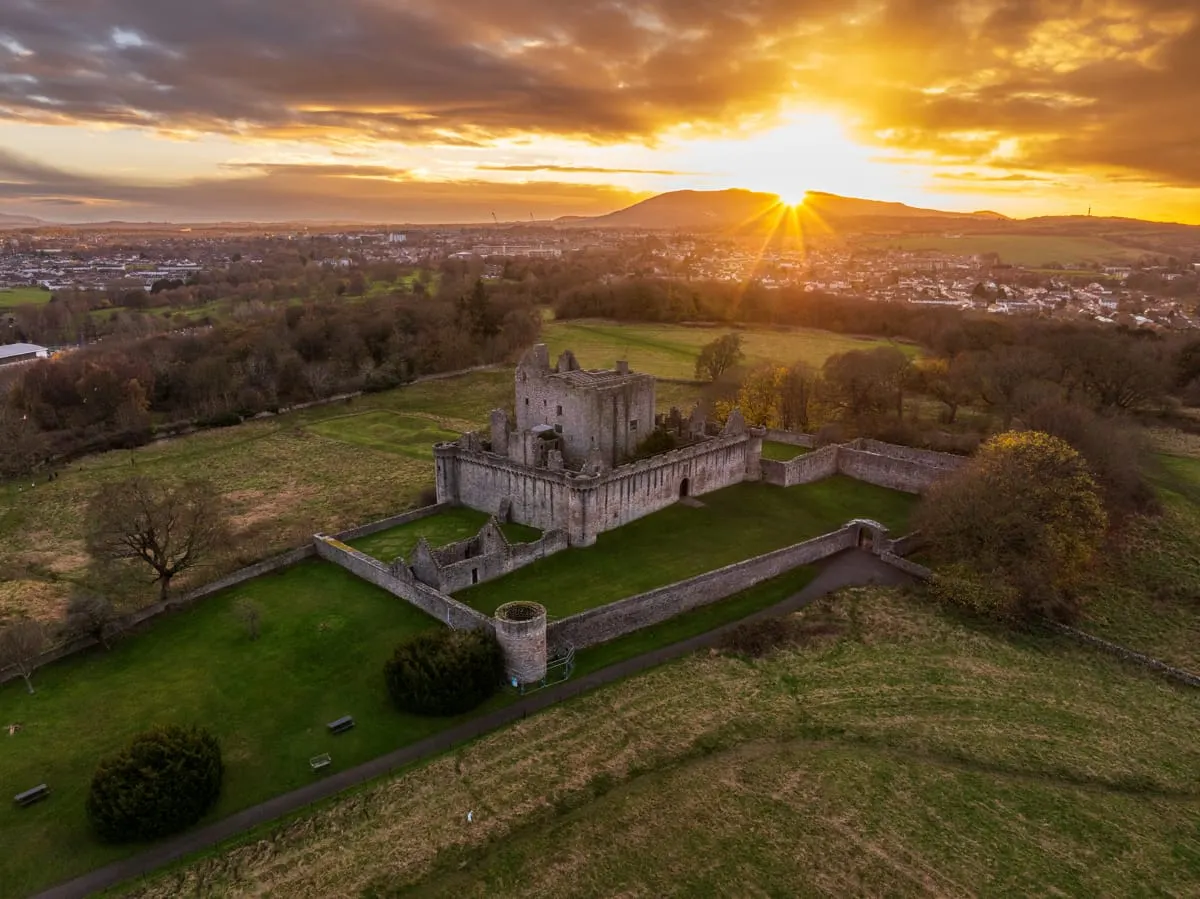 Aerial view of a ruined medieval castle with defensive walls at sunset, surrounded by green fields and trees with a town visible in the distance.