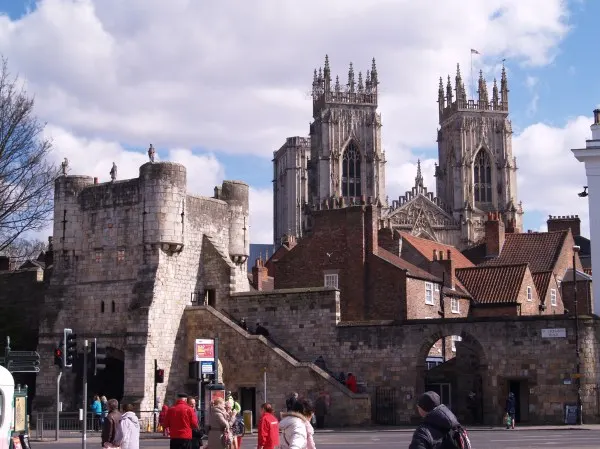 Medieval stone gatehouse with Gothic cathedral towers behind, visitors gathering in courtyard.