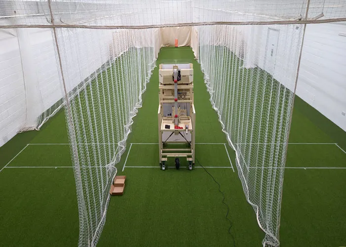 Indoor batting cage with artificial grass, netting dividers, and a bowling machine down the centre alley.
