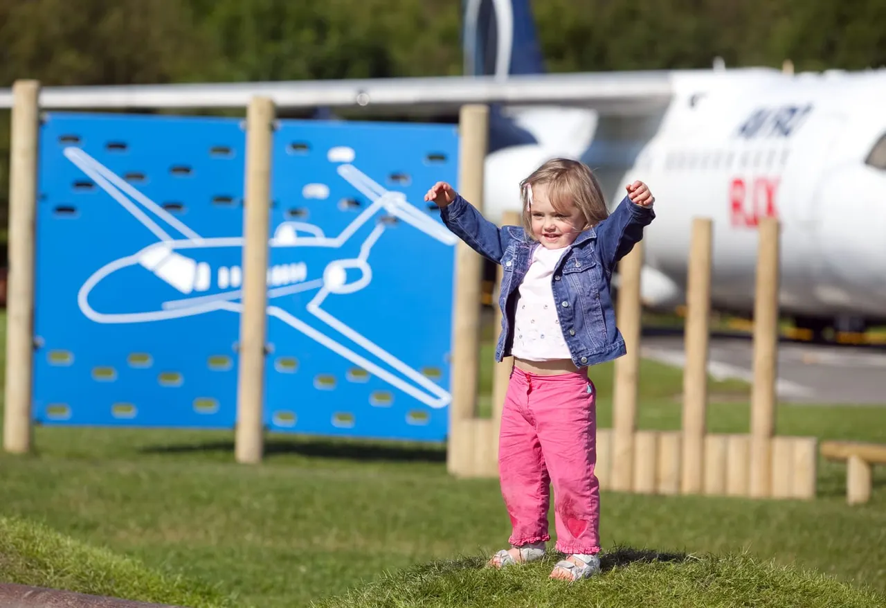Toddler with arms raised celebrating on grass with aeroplane-themed playground panels and aircraft behind.