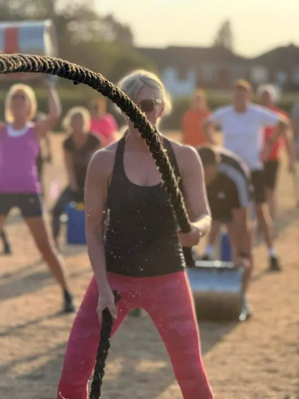 Woman swinging a thick rope during an outdoor fitness class with other participants watching in the background.