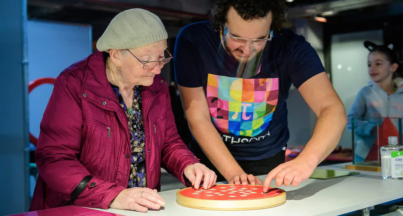 An older woman and a younger man work together on an interactive maths activity involving a red circular board with white dots at an indoor science centre.